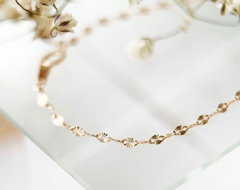 May include: A delicate gold chain bracelet featuring a series of small, flat, oval-shaped links with a textured, sunburst-like pattern on each segment. The chain is thin and reflective, showcasing a polished metallic finish throughout the design.