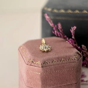 May include: A gold pendant featuring a central clear stone surrounded by a cluster of smaller clear stones in a floral arrangement. The pendant rests on a dusty rose velvet hexagonal box with gold trim, positioned near a dark velvet box.