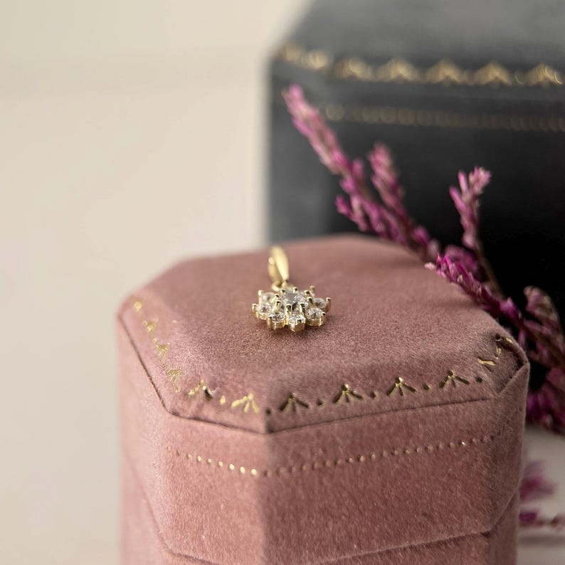 May include: A gold pendant featuring a central clear stone surrounded by a cluster of smaller clear stones in a floral arrangement. The pendant rests on a dusty rose velvet hexagonal box with gold trim, positioned near a dark velvet box.