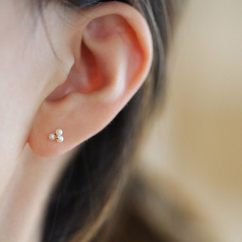 May include: A close-up of a gold stud earring featuring three tiny, white pearls in a cluster design. The earring is set against the backdrop of a person's ear, highlighting the jewellery's delicate nature.