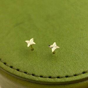 May include: A pair of small, gold-colored star-shaped stud earrings. The earrings are set against a green background. The stars have a textured surface and are likely made of metal.