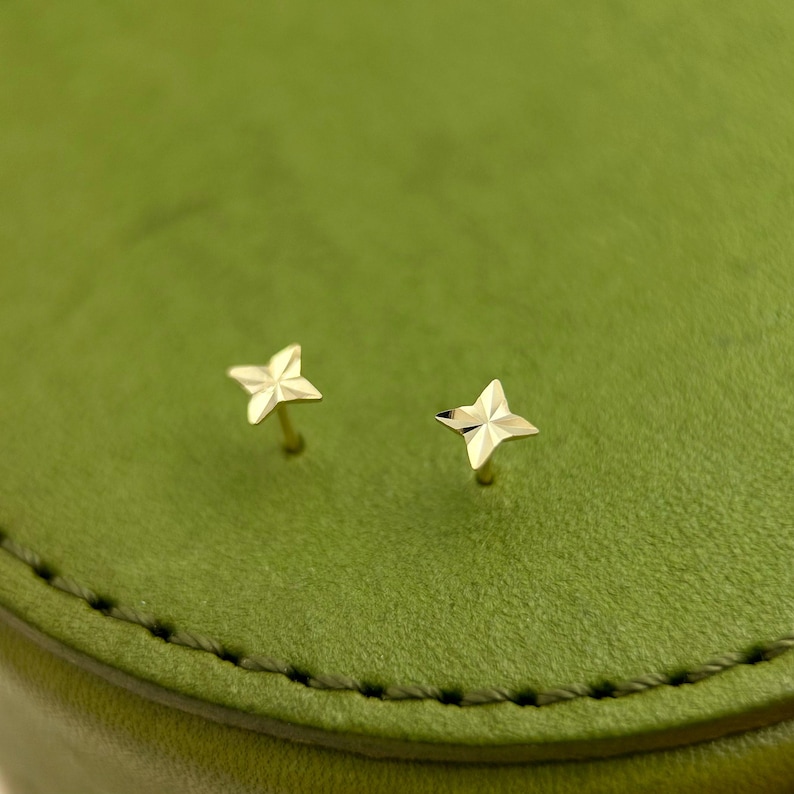 May include: A pair of small, gold-colored star-shaped stud earrings. The earrings are set against a green background. The stars have a textured surface and are likely made of metal.