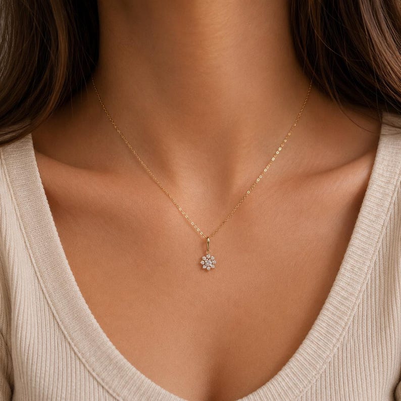 May include: A delicate gold chain necklace featuring a small, circular pendant shaped like a flower. The pendant is set with a central clear gemstone surrounded by a ring of smaller, sparkling clear stones, creating a floral design.