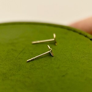 May include: A pair of small, gold-colored stud earrings. Each earring features a delicate, stylized star design. The earrings are set against a green background, highlighting their metallic sheen. The earrings are a jewelry item.