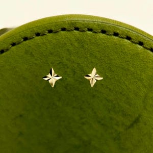 May include: A pair of gold-colored star-shaped stud earrings. The earrings are displayed against a textured, green background. The earrings have a four-pointed star design, with each point having a faceted appearance.