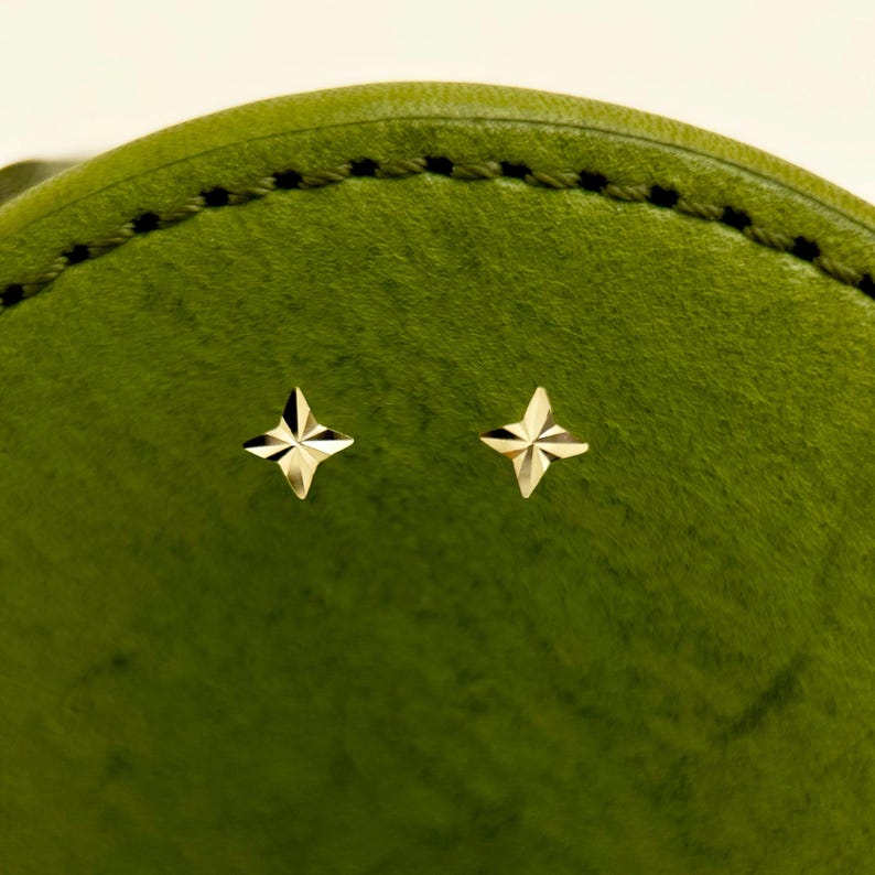 May include: A pair of gold-colored star-shaped stud earrings. The earrings are displayed against a textured, green background. The earrings have a four-pointed star design, with each point having a faceted appearance.