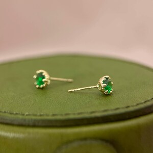 Puede incluir: Un par de pendientes de bot&oacute;n de oro con piedras preciosas verdes. Los pendientes tienen un dise&ntilde;o sencillo con una peque&ntilde;a piedra preciosa redonda engastada en un engaste de oro.