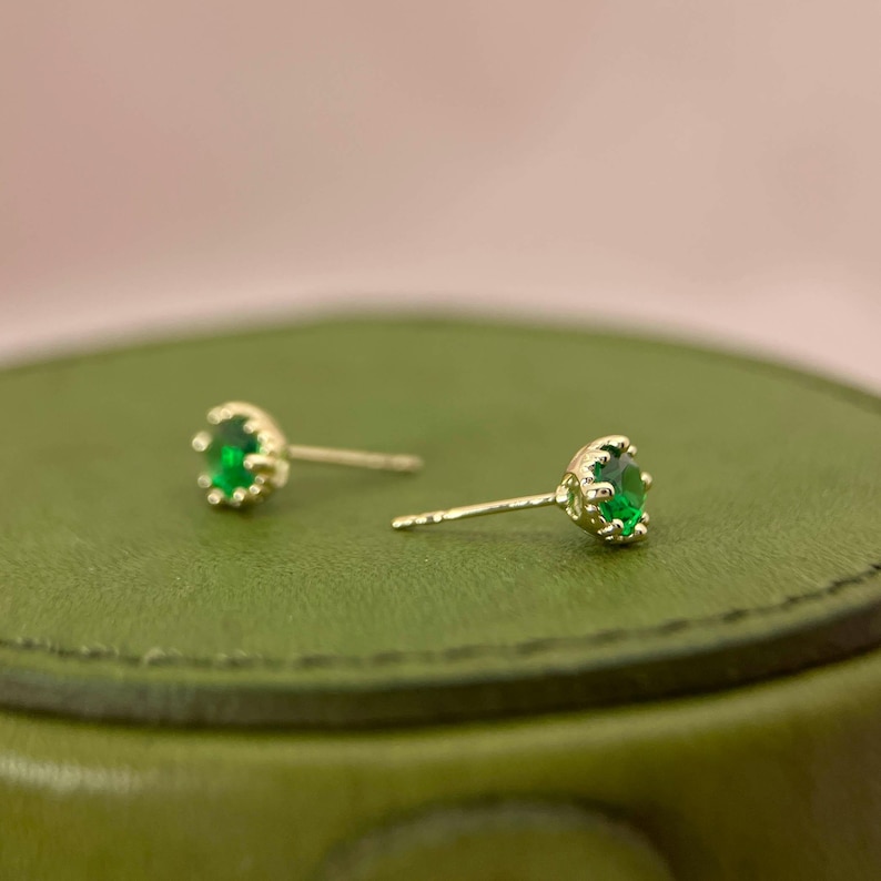 Puede incluir: Un par de pendientes de bot&oacute;n de oro con piedras preciosas verdes. Los pendientes tienen un dise&ntilde;o sencillo con una peque&ntilde;a piedra preciosa redonda engastada en un engaste de oro.