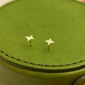 May include: A pair of small, gold-colored star-shaped stud earrings. The earrings are on a green surface. The studs have a four-point star design and are likely made of metal.