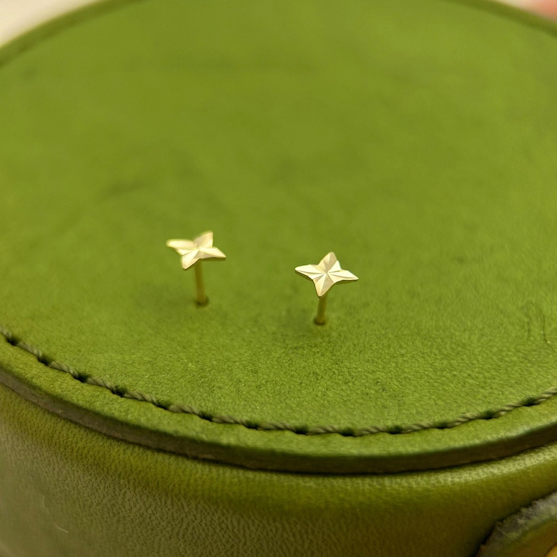 May include: A pair of small, gold-colored star-shaped stud earrings. The earrings are on a green surface. The studs have a four-point star design and are likely made of metal.