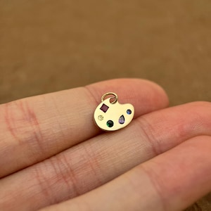 Op de afbeelding: Een gouden kunstenaars-paletbedel met gekleurde edelstenen. De bedel heeft een rode, groene, blauwe en gele edelsteen.