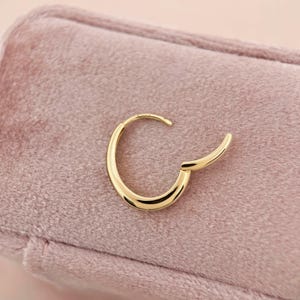 May include: A gold hoop earring with a smooth, rounded design. The earring is on a pink velvet surface.