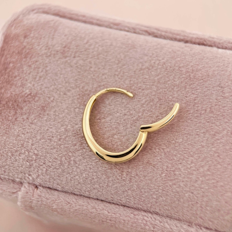 May include: A gold hoop earring with a smooth, rounded design. The earring is on a pink velvet surface.