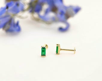 Puede incluir: Dos pendientes de botón dorados con piedras preciosas rectangulares de color verde esmeralda. Los pendientes se muestran sobre una superficie blanca, con flores azules borrosas en el fondo. Los pendientes tienen un poste y una parte trasera plana.