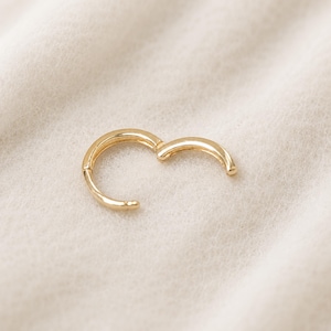 May include: A gold-colored, hinged hoop earring, open and ready to be clasped. The earring is set against a soft, off-white background. The earring's design is simple and elegant, with a smooth, polished finish.