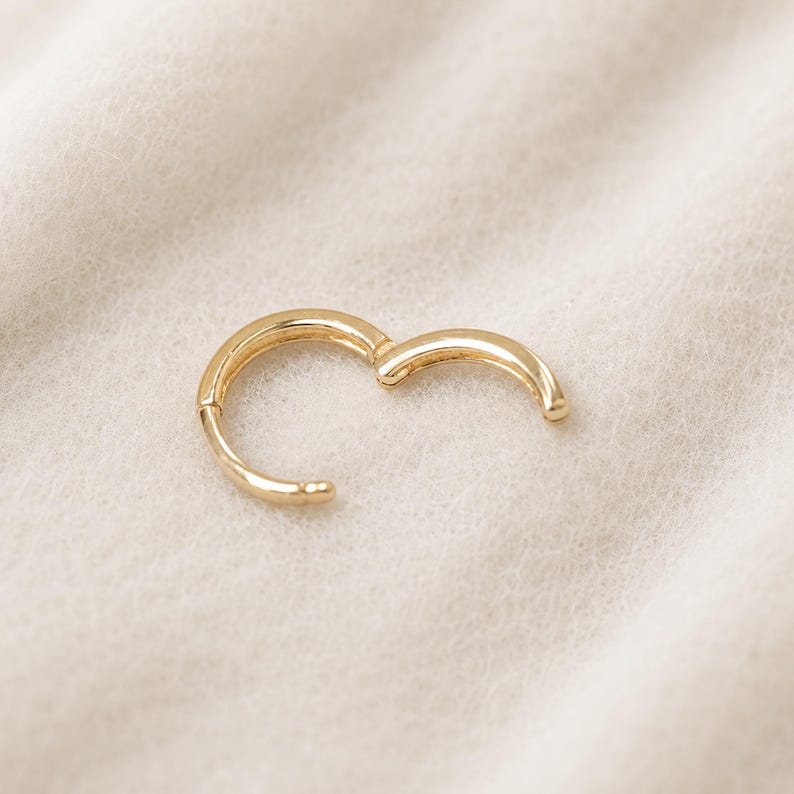 May include: A gold-colored, hinged hoop earring, open and ready to be clasped. The earring is set against a soft, off-white background. The earring's design is simple and elegant, with a smooth, polished finish.
