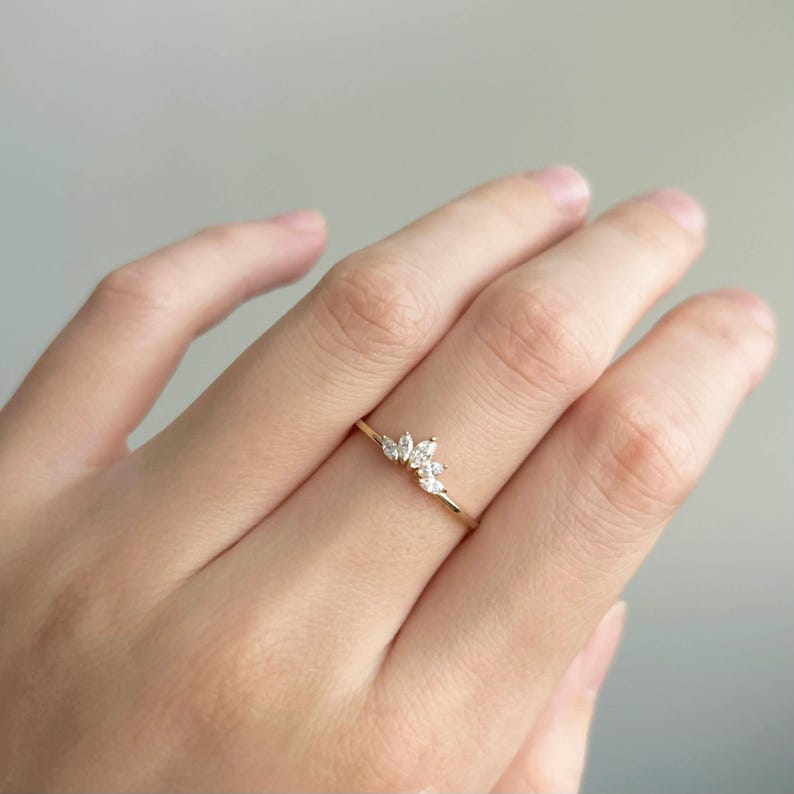 Puede incluir: Un delicado anillo de oro con un grupo de peque&ntilde;as piedras preciosas transparentes. La banda del anillo es fina y sencilla, con las piedras preciosas dispuestas en un patr&oacute;n floral o de hoja. El anillo se muestra en un dedo.