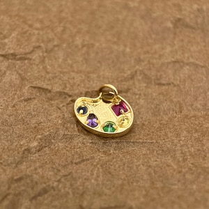 Op de afbeelding: Gouden kunstenaars-paletbedel met gekleurde edelstenen. De bedel heeft een paarse, groene, rode en blauwe edelsteen.