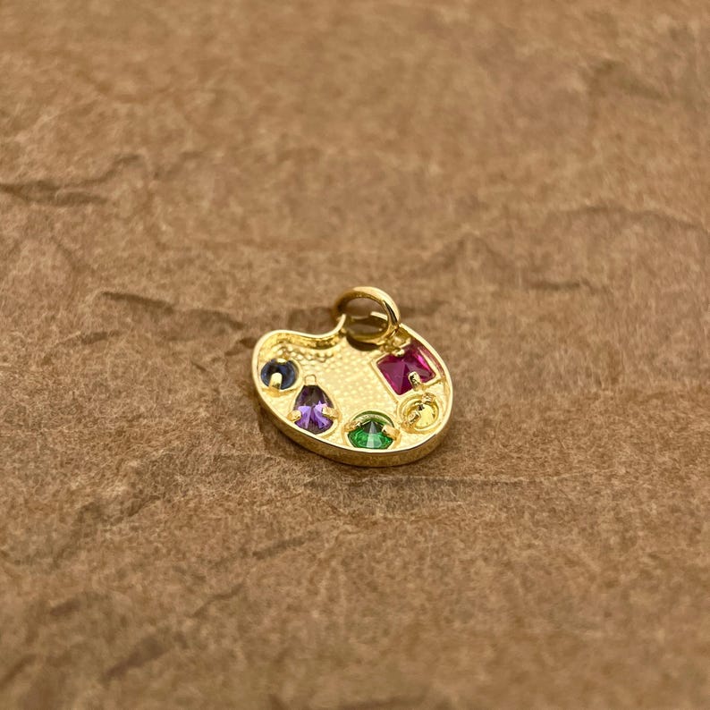 Op de afbeelding: Gouden kunstenaars-paletbedel met gekleurde edelstenen. De bedel heeft een paarse, groene, rode en blauwe edelsteen.