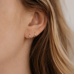 May include: Close-up of a person's ear with two small, gold hoop earrings. The earrings are simple and elegant, with a polished finish. The skin tone is fair, and the hair is blonde.