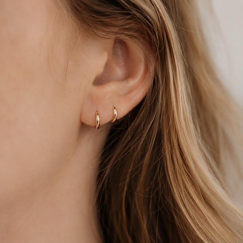 May include: Close-up of a person's ear with two small, gold hoop earrings. The earrings are simple and elegant, with a polished finish. The skin tone is fair, and the hair is blonde.