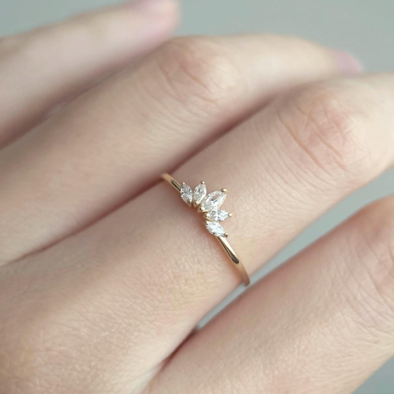 Puede incluir: Un delicado anillo de oro con un dise&ntilde;o curvo adornado con varias piedras preciosas peque&ntilde;as y claras. La fina banda pulida del anillo sostiene un grupo de piedras preciosas, creando un brillo sutil. El anillo se muestra en un dedo.