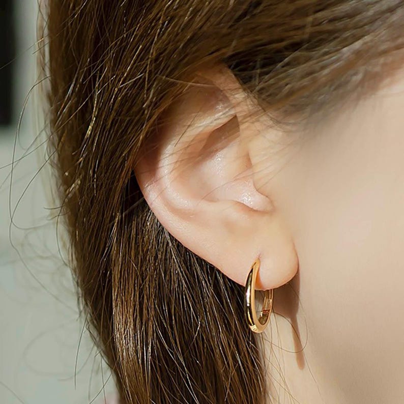 May include: A close-up of a person's ear with a gold hoop earring. The earring is simple and elegant, and it is a great choice for everyday wear.