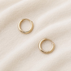 May include: Two gold-colored hoop earrings. The earrings are circular with a hinged closure. The earrings are displayed on a cream-colored, textured surface. The earrings are a simple, classic design.