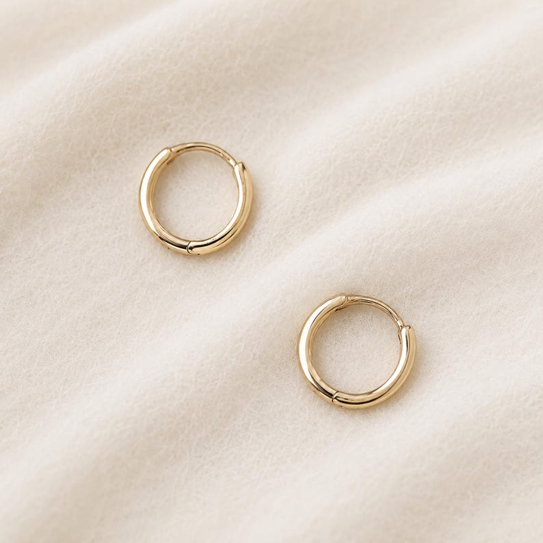 May include: Two gold-colored hoop earrings. The earrings are circular with a hinged closure. The earrings are displayed on a cream-colored, textured surface. The earrings are a simple, classic design.