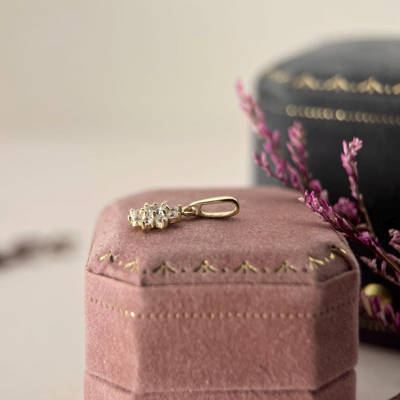 May include: A gold pendant featuring a cluster of small, clear, round-cut stones arranged in a floral pattern. The pendant includes a simple, polished gold bail at the top for attachment to a chain, showcasing a bright, reflective metallic finish.