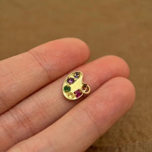 Op de afbeelding: Gouden charme in de vorm van een schilderspalet met vijf gekleurde edelstenen. De charme heeft een lus om aan een ketting of armband te bevestigen.