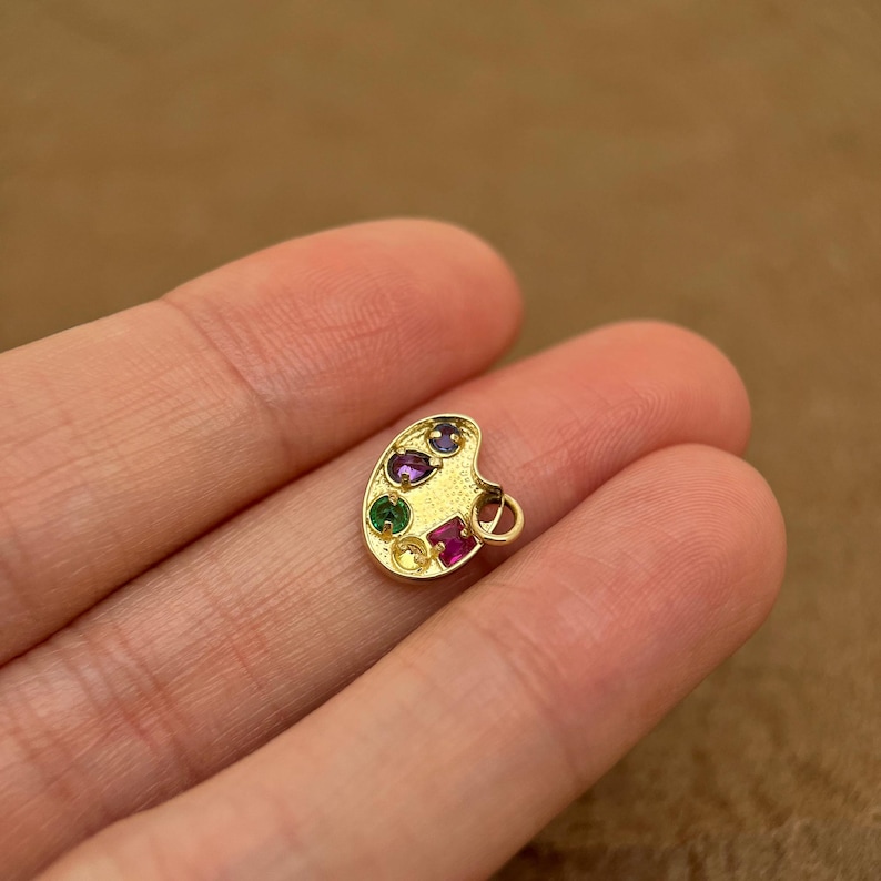 Op de afbeelding: Gouden charme in de vorm van een schilderspalet met vijf gekleurde edelstenen. De charme heeft een lus om aan een ketting of armband te bevestigen.