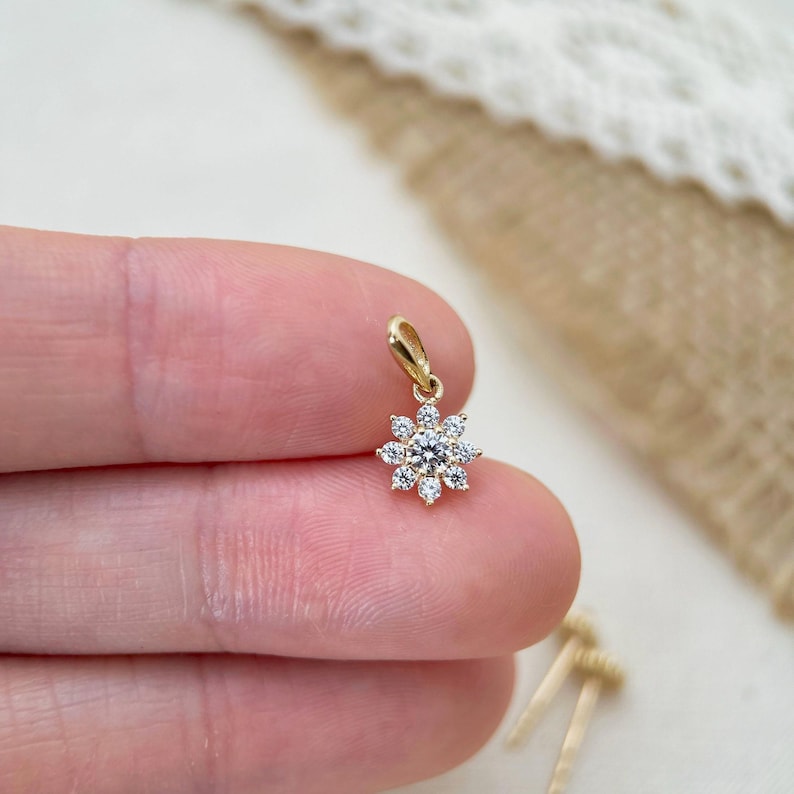 May include: A small gold pendant featuring a central round brilliant-cut diamond surrounded by eight smaller round diamonds arranged in a floral starburst pattern, complete with a simple gold bail at the top for attachment to a necklace chain.