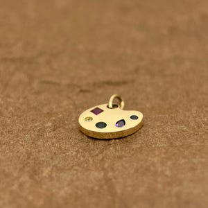 Op de afbeelding: Een gouden kunstenaars-paletbedel met gekleurde edelstenen. De bedel is gevormd als een traditioneel kunstenaars-palet met een gat aan de bovenkant om aan een ketting of armband te bevestigen.