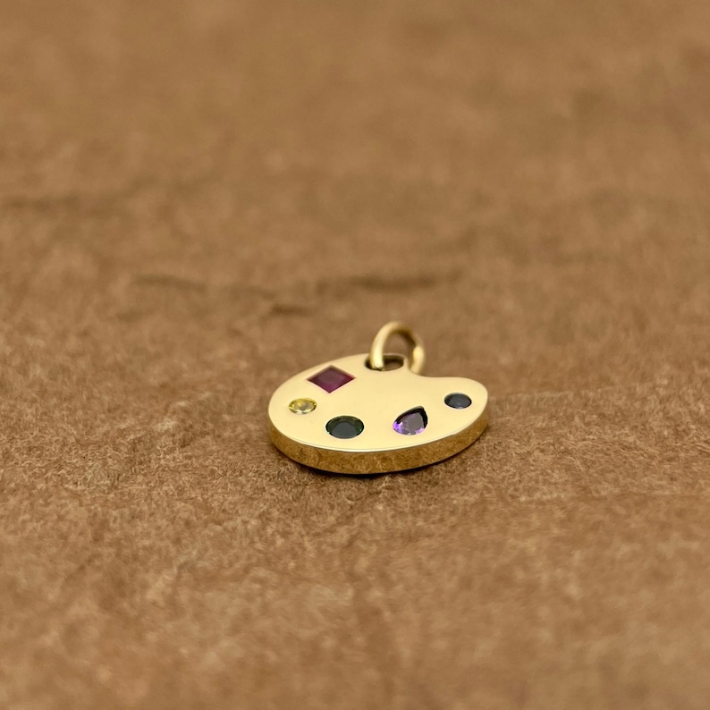 Op de afbeelding: Een gouden kunstenaars-paletbedel met gekleurde edelstenen. De bedel is gevormd als een traditioneel kunstenaars-palet met een gat aan de bovenkant om aan een ketting of armband te bevestigen.
