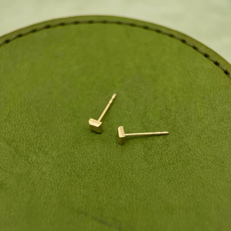 May include: A pair of gold square stud earrings on a green surface.