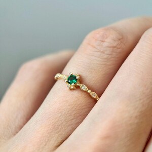 Puede incluir: Un delicado anillo de oro con una peque&ntilde;a piedra preciosa redonda de color verde esmeralda engastada en una montura de garras. La banda est&aacute; adornada con peque&ntilde;os diamantes brillantes.