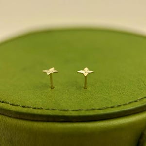 May include: A pair of small, gold-colored stud earrings. Each earring features a four-petal flower design. The earrings are set against a green background.