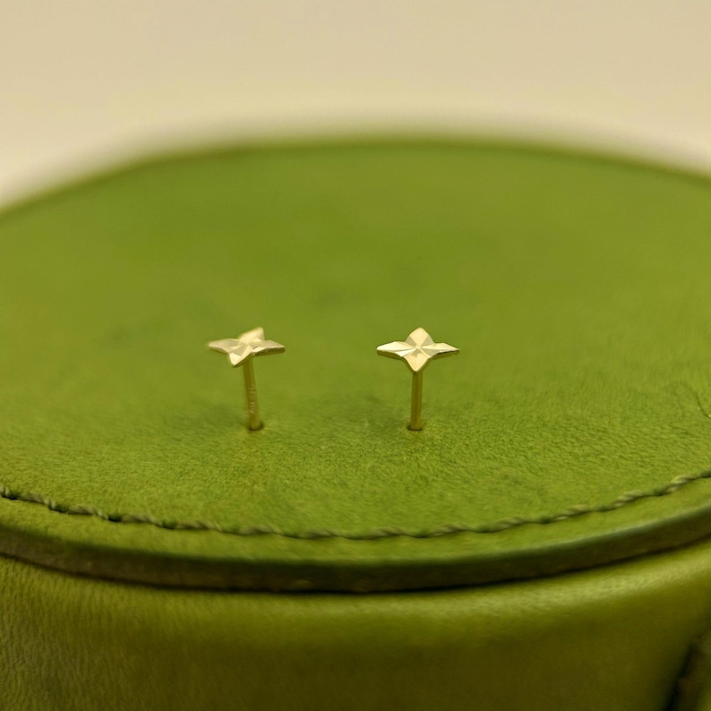 May include: A pair of small, gold-colored stud earrings. Each earring features a four-petal flower design. The earrings are set against a green background.