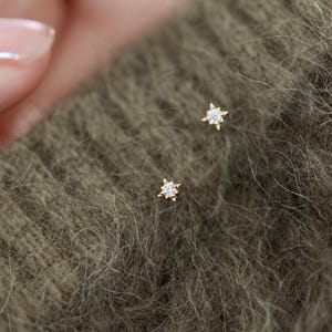 Pode incluir: Um par de pequenos brincos de ouro, cada um com uma pedra clara central cravada numa armação em forma de estrela de quatro pontas. Os brincos estão colocados sobre um tecido de malha verde-azeitona texturado, com um design delicado.