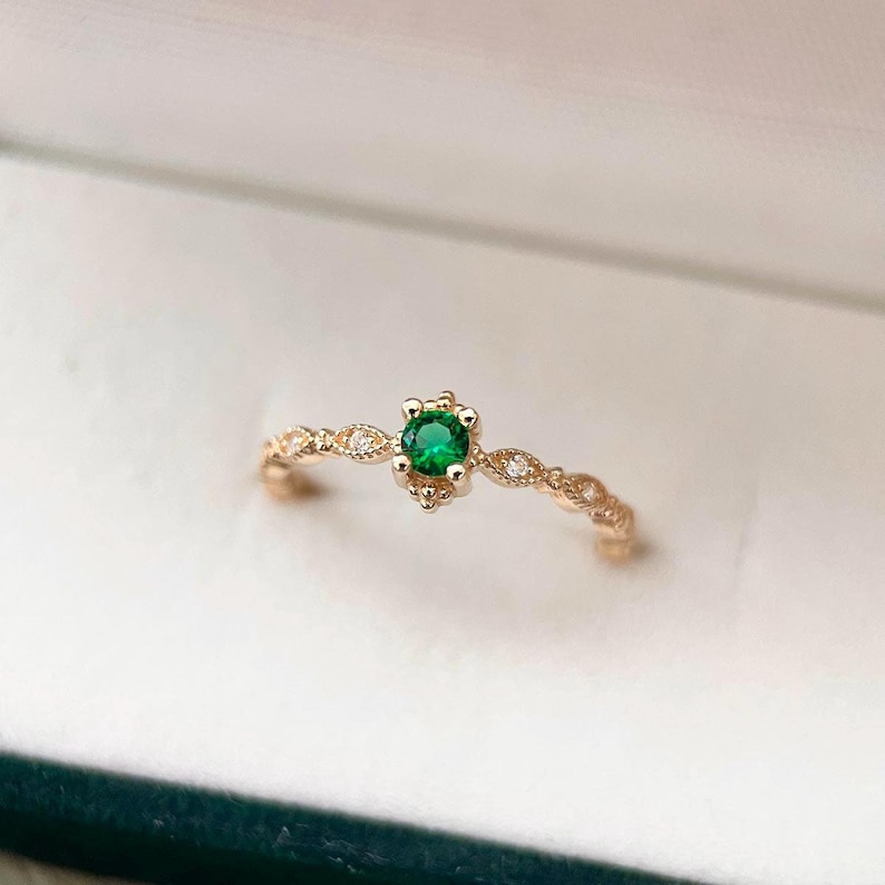 Puede incluir: Un delicado anillo de oro con una peque&ntilde;a piedra preciosa verde redonda rodeada de diminutos diamantes. El anillo tiene un dise&ntilde;o delicado y ornamentado.