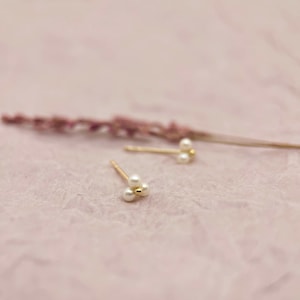 May include: Gold-coloured stud earrings, each with three small, white pearls in a cluster. The earrings are set against a soft pink background, with a dried lavender stem in the background.