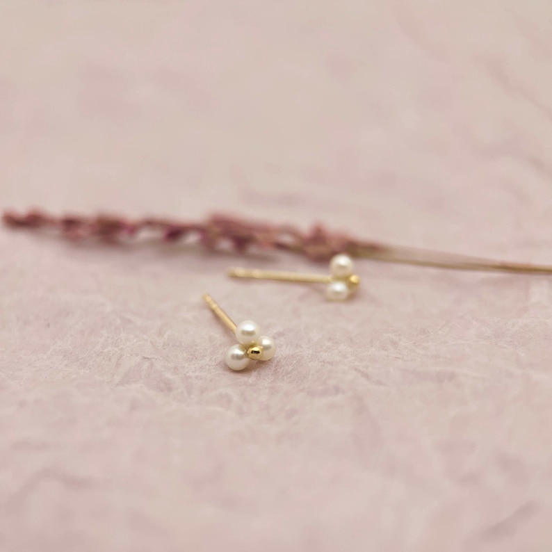 May include: Gold-coloured stud earrings, each with three small, white pearls in a cluster. The earrings are set against a soft pink background, with a dried lavender stem in the background.