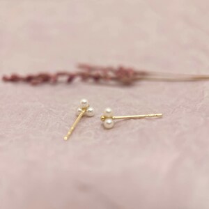 May include: Two gold stud earrings, each with three small, round pearls. The earrings are set against a soft pink background, with a blurred sprig of lavender in the background. The earrings have a delicate and elegant design.
