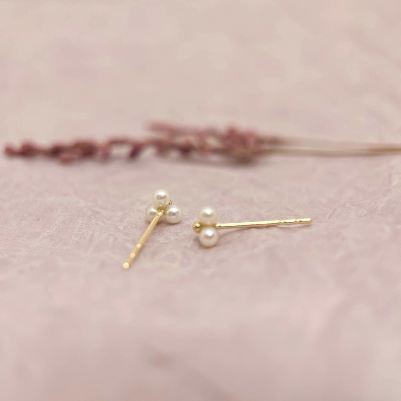 May include: Two gold stud earrings, each with three small, round pearls. The earrings are set against a soft pink background, with a blurred sprig of lavender in the background. The earrings have a delicate and elegant design.