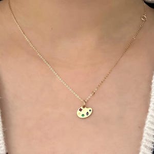 Op de afbeelding: Een gouden ketting met een klein gouden hanger in de vorm van een schilderspalet. Het palet heeft vijf gekleurde edelstenen: rood, groen, blauw, geel en paars.