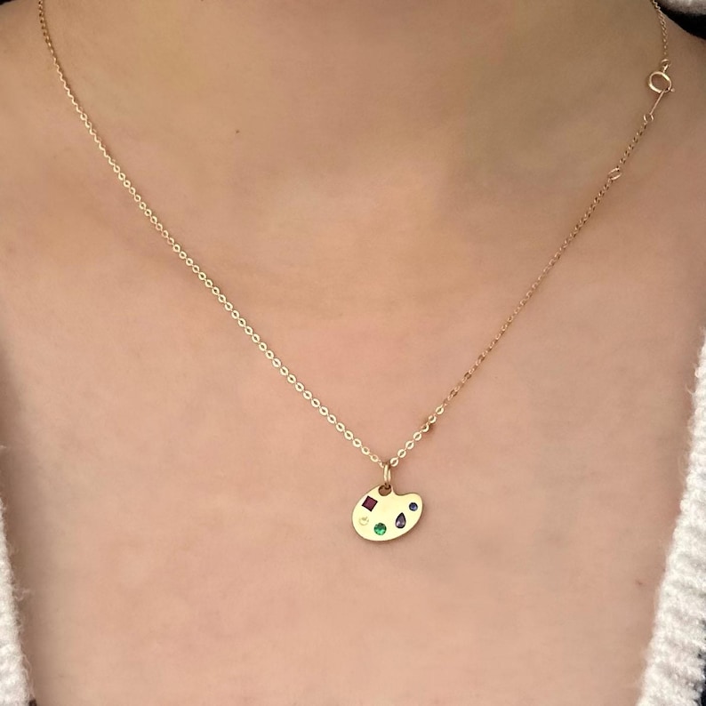 Op de afbeelding: Een gouden ketting met een klein gouden hanger in de vorm van een schilderspalet. Het palet heeft vijf gekleurde edelstenen: rood, groen, blauw, geel en paars.