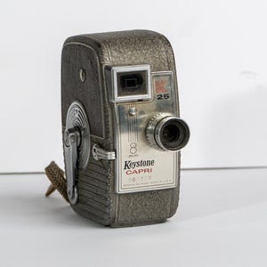 May include: A silver Keystone Capri 8 mm movie camera with a black leather strap. The camera has a lens cap and a red "K" logo on the side.