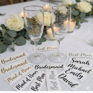 Könnte beinhalten: Zwei Sektgläser mit "Bride" und "Groom" in goldener und silberner Schrift. Mehrere Tischkarten mit Namen und Titeln wie "Bridesmaid", "Best Man" und individuellen Namen sind auf einer weißen Tischdecke angeordnet, mit Blumen- und Kerzendekorationen.