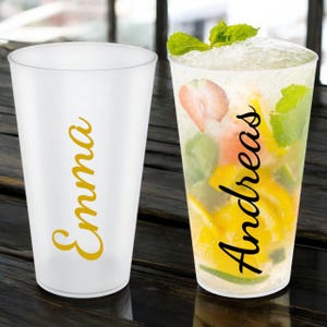 Könnte beinhalten: Zwei transparente Plastikbecher, einer leer und der andere mit einem erfrischenden Getränk gefüllt. Der leere Becher trägt den Namen "Emma" in goldener Schrift. Der gefüllte Becher enthält Eis, Früchte und Minze, mit dem Namen "Andreas" in goldener Schrift.